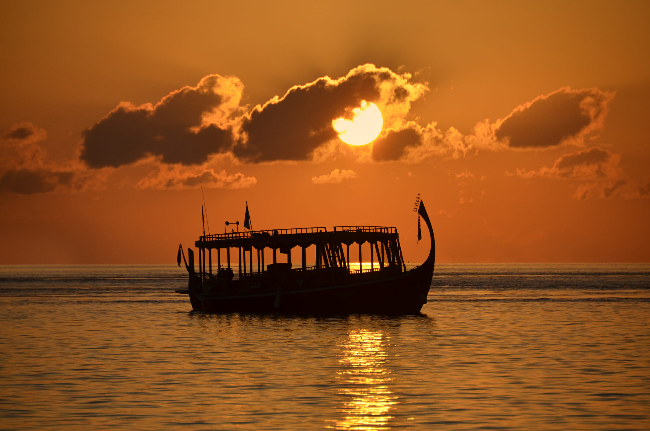 Silhouette of a Dhoni (traditional Maldivian boat) in the tropical lagoon of Biyadhoo Island, Maldives, at sunset with bright orange-reddish sky.